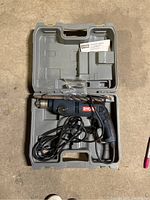 Ryobi hammer drill with power cord inside an open gray plastic carrying case with compartments. Case shows signs of wear and latch does not lock.