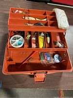 Orange plastic layered tackle box filled with various fishing lures, hooks, baits, and fishing line