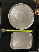 Both round and rectangular stainless steel serving trays shown side by side on a black surface with measuring tape indicating sizes.