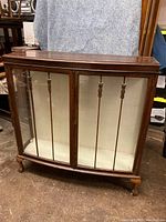 Front view showing wood frame, bow front curved glass doors with vertical wooden dividers, interior white fabric lining, and two cabinet doors.