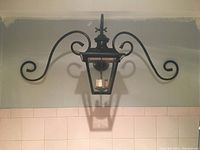 Photo showing the black metal exterior light fixture with classic lantern shape, clear glass panels, and wrought iron scrollwork arms mounted on a beige wall.