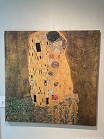 Photo of a square canvas print depicting Gustav Klimt's The Kiss showing a couple in an embrace with richly decorated gold and patterned robes and a floral base.