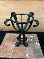 Black wrought iron flower pot holder with four curled legs, round opening with decorative curved extensions, placed on a marble surface against a wood background.