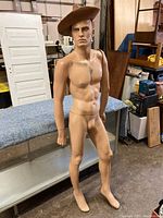 Full view of the vintage male mannequin located indoors on a grey table with miscellaneous background items. The mannequin has realistic facial features and two right arms, standing without a hat.