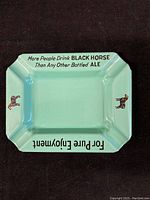 Top-down image of a light green glazed porcelain ashtray with Black Horse Ale advertising text and horse illustrations.