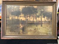 Framed vintage print showing a landscape with trees, a water feature with a figure near it, muted colors, protected by glass with visible wear on frame.
