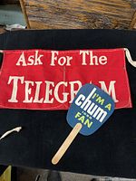 Photo of a red telegram apron with white text 'Ask For The TELEGRAM' and a blue handheld fan with 'I'm A chum 1050 FAN' text.