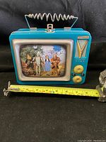 Front view showing overall tin lunchbox with Wizard of Oz character graphics on screen, spiral antenna handle, and size reference with measuring tape.