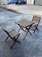 Three-piece teak bistro set including square slatted table and two folding chairs displayed on asphalt