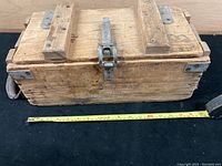 Closed view of wooden WW II grenade/ammunition box showing locked metal latch, metal corner brackets and wood construction with wear and patina. Tape measure placed for scale at front showing approx 18 inches length.