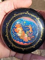 Hand held close-up of the antique round black lacquer box showing the detailed painted scene of a troika with three horses and driver, gold decorative border, and some visible crazing on the lacquer surface.