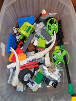 Top-down view of translucent plastic container with assorted LEGO pieces inside, showing variety of colors and types of LEGO bricks and parts.