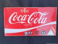 Photo of a red and white vintage Coca Cola plexiglass sign with hockey team stickers along the bottom edge, showing the 'Enjoy Coca Cola' slogan.