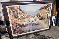 Large framed picture of a European cityscape placed outdoors showing details of buildings, tables and chairs, mountains, and a waterfront.