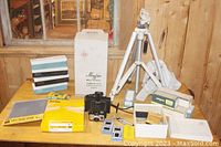 Overview of lot showing vintage cameras, aluminum tripod, boxes of slides, photographic grey card and film rolls on wooden table