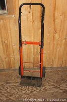 Metal dolly with black handle, orange frame, and two wheels, standing upright against wooden wall.