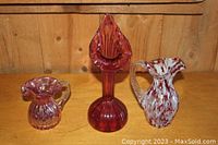 Three blown glass pieces on wooden surface: two pitchers and one vase, all in red glass with artistic details