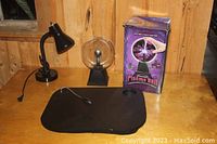 Photo showing one plasma ball on black base next to original box, smaller plasma ball on black base, black adjustable office lamp with cord, all arranged on wooden table.