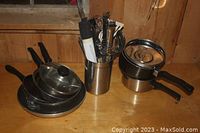 Photo showing three stainless steel pots with black handles and glass lids, two frying pans, and a cylinder utensil holder containing various metal kitchen utensils.