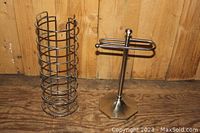 Both metal toilet paper holders against a wooden backdrop on a wooden floor. One holder is a vertical cylindrical rack; the other is a stand with dual arms and hexagonal base.