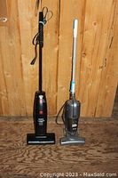 Two upright vacuum cleaners side by side against a wooden backdrop, left one black Eureka The Boss Lite, right one silver Shark.