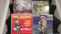 Four vinyl records including two albums featuring Hank Williams Sr and one with Hank Williams Jr, displayed on a black cloth background.