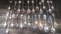 Photo showing all 26 souvenir silver spoons arranged in two rows on a dark wooden surface. Spoons have various emblems, crests, and colored enamel on handle tops.