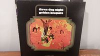 Front cover of the Three Dog Night 'Golden Bisquits' vinyl record album showing band members under a tree with a colorful border on a black background.