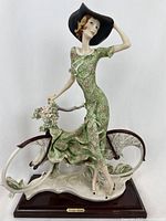 Front view of the Girl with Bicycle figurine showing the elaborate floral-patterned green dress and wide-brimmed black hat, mounted on wooden base.