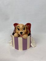 Front view of Lady figurine lying in purple and cream striped box with large red bow.