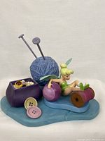 Front view of Tinker Bell sculpture with sewing-themed items on a blue base