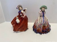 Front view of two Royal Doulton bone china figurines Autumn Breezes (red dress) and Easter Day (multi-colored dress).