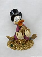 Oblique front view of Scrooge McDuck figurine seated on pile of gold-colored coins holding stacks of coins.