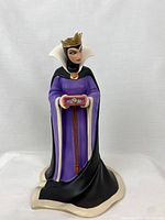 Front view of the Evil Queen figurine, showing her holding a red box with a heart and wearing a purple and black dress with white accents and a gold crown.