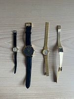 Four women's watches laid flat on wooden surface - two with black leather straps and two with gold-tone metal bands.