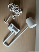 White Ikea Nymane adjustable wall lamp with cylindrical shade, adjustable arm, wall mount base, power cord and rotary on/off switch, shown on wooden surface.