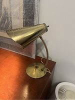 Side angle of a brass Mid-Century Modern style desk lamp on wooden surface, showing the conical lampshade and flexible neck with visible wear and patchy patina on the brass surface.