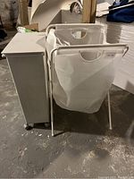 Photo showing a white rectangular Muji garbage can with caster wheels next to a white Ikea fabric hamper with a metal frame and handles, positioned on a concrete floor with various background objects.