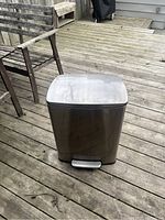 Front view of the stainless steel Simplehuman garbage can on a wooden deck, showing the foot pedal and top lid.