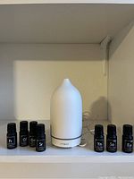 Image shows a white Vitruvi ceramic essential oil diffuser and seven black-topped small essential oil bottles arranged on a white shelf.
