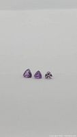 Three natural purple amethyst gemstones, trillion cut, displayed in a row on a light background.