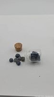 Glass vial containing natural blue sapphire crystals with cork stopper beside it and crystals scattered outside