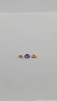 Three natural gemstones on plain background: One oval purple amethyst in center, two pear-shaped golden citrine on sides.