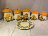Five ceramic jars with raised mushroom motifs and orange mushroom cap lids, arranged in a row with matching ceramic mushroom-themed plate in front.