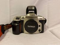Front view of Nikon F60 camera body with Tamron lens cap and pop-up flash raised. Shows silver camera body with black grip and orange accent on grip.