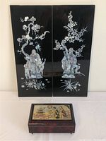 Front view of the pair of mother of pearl lacquered wall art panels with Asian figures and flora, leaning against the wall behind the jewelry box.