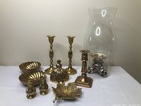 Photo showing entire lot of brass decor items arranged on a table including candle holders, dishes, bell, and hurricane lantern.