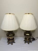 Pair of matching brass base table lamps with white pleated fabric shades on a white cloth surface against a white wall.