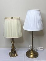 Two lamps with brass bases, one with a tall slim round base and white pleated shade, the other with a shorter ornate base and off-white pleated shade.
