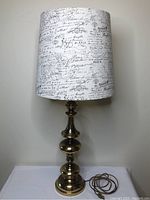 Full view of the large brass table lamp standing on a white surface, showing the curvy polished brass base and connected power cord.